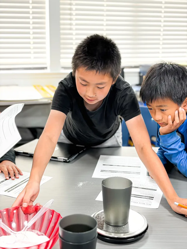 Students work with a bowl of sugar, cups, worksheets, and a digital scale