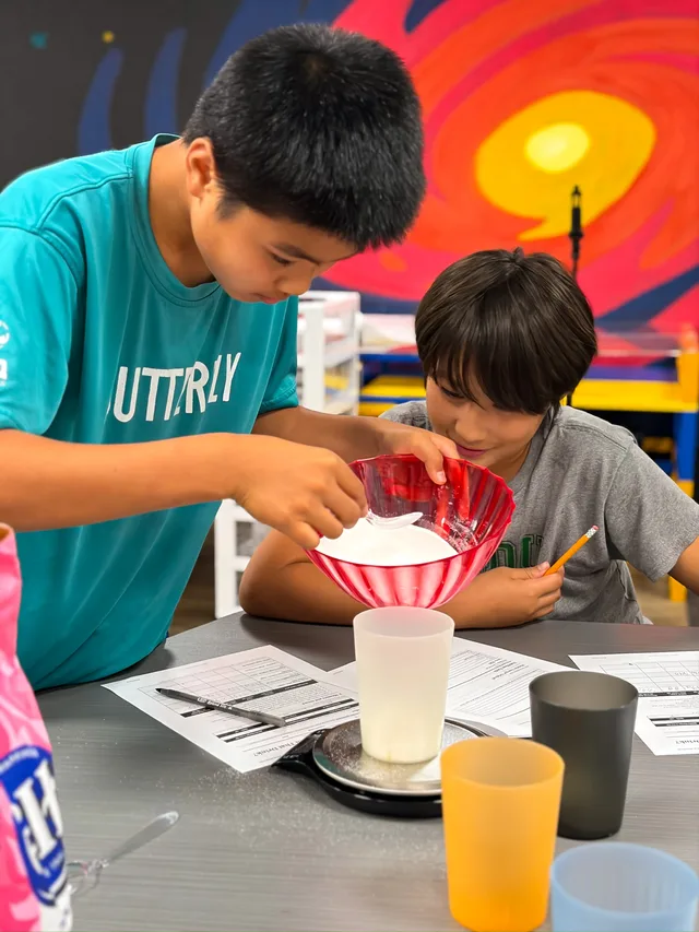 Two students scoop sugar from a red bowl into a cup on a scale